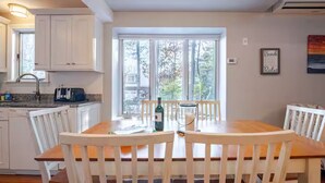 Comedor con mesa y sillas de madera, ventana con vista a árboles y muebles de cocina blancos.