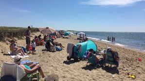Una playa con gente relajándose bajo sombrillas, algunos sentados en sillas y unos pocos caminando por la orilla.