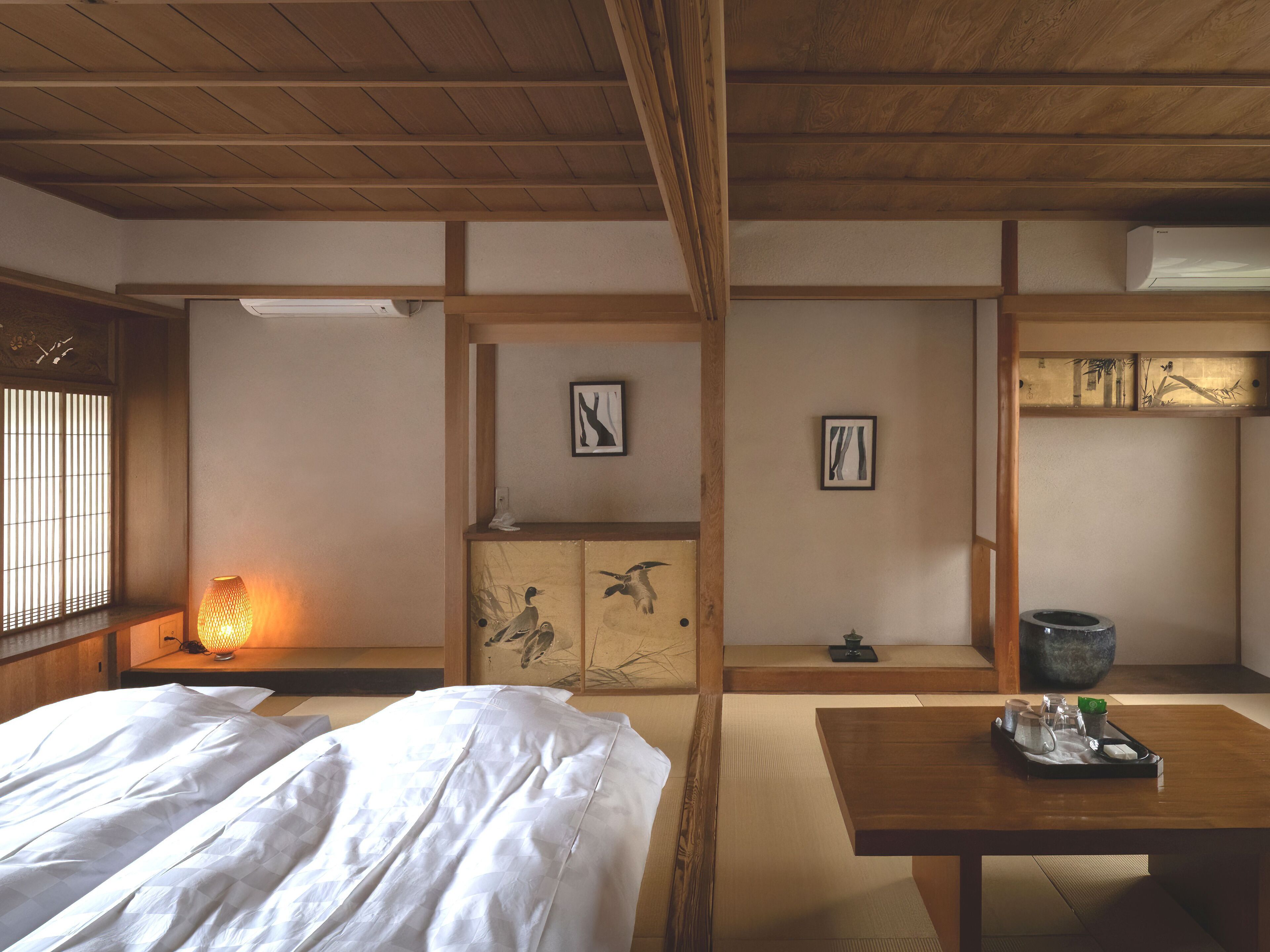 A traditional Japanese room with tatami flooring, a low wooden table with a tray, a bed with white bedding, and a window with vertical blinds.