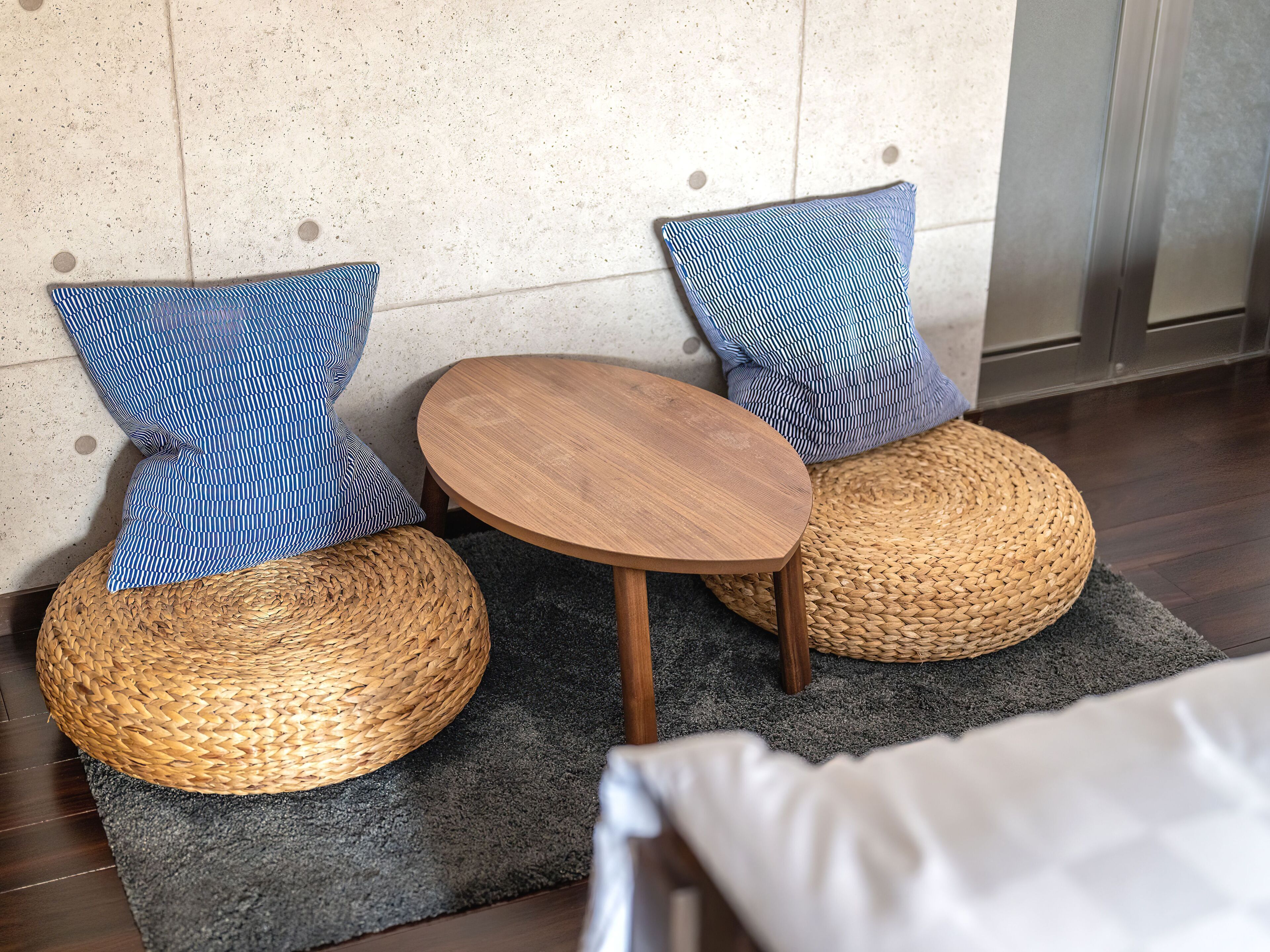Un petit coin salon avec deux poufs en fibres tressées et une table ronde en bois, le tout placé devant un mur à texture et sur un tapis foncé.
