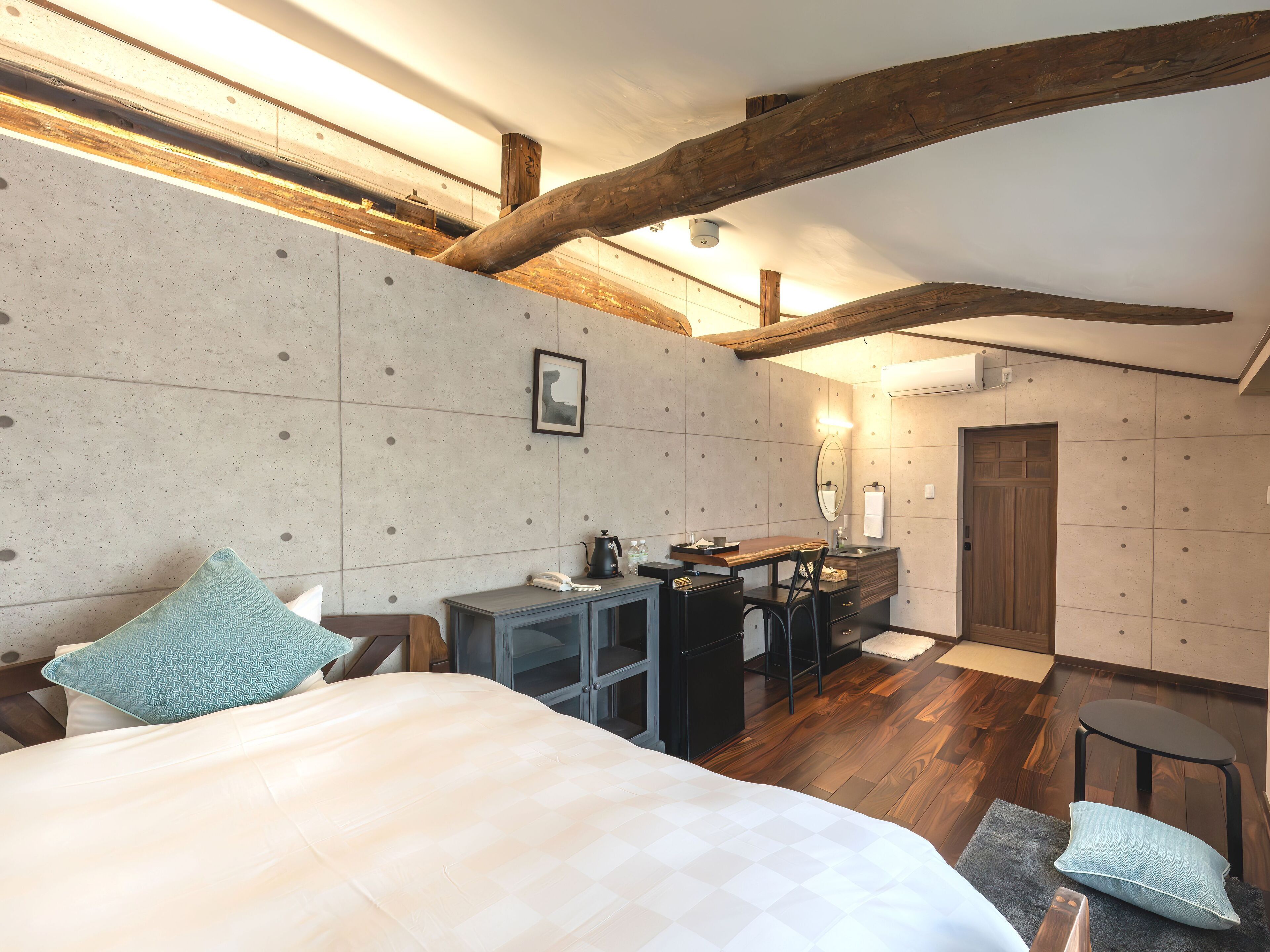 A bedroom with a bed, a desk, and a chair. There is a wooden ceiling beam and a wall-mounted light fixture.