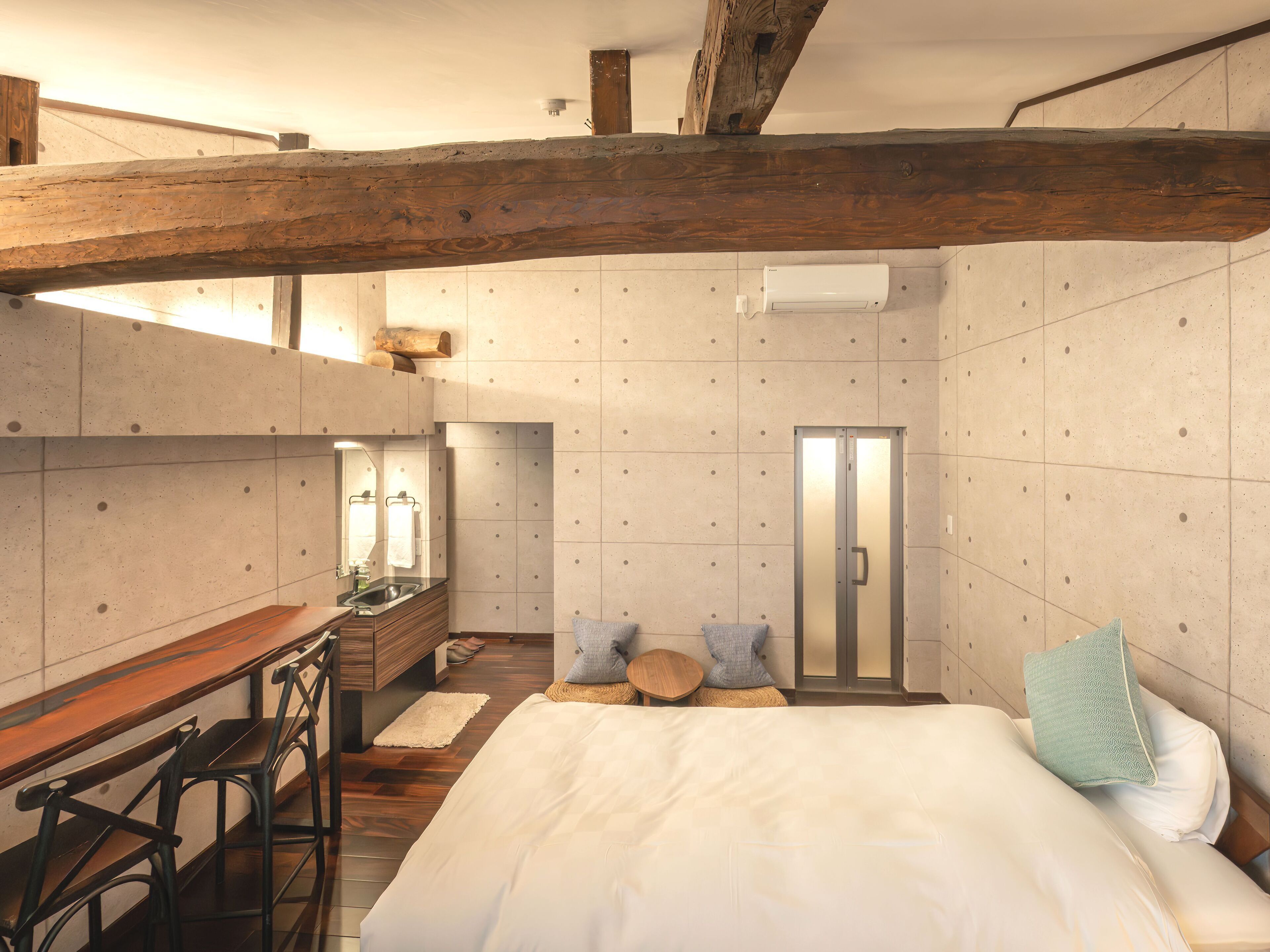 Un dormitorio con una cama grande, un escritorio de madera y una silla. Hay una pequeña mesa con un taburete, un espejo y una toalla colgada en la pared.