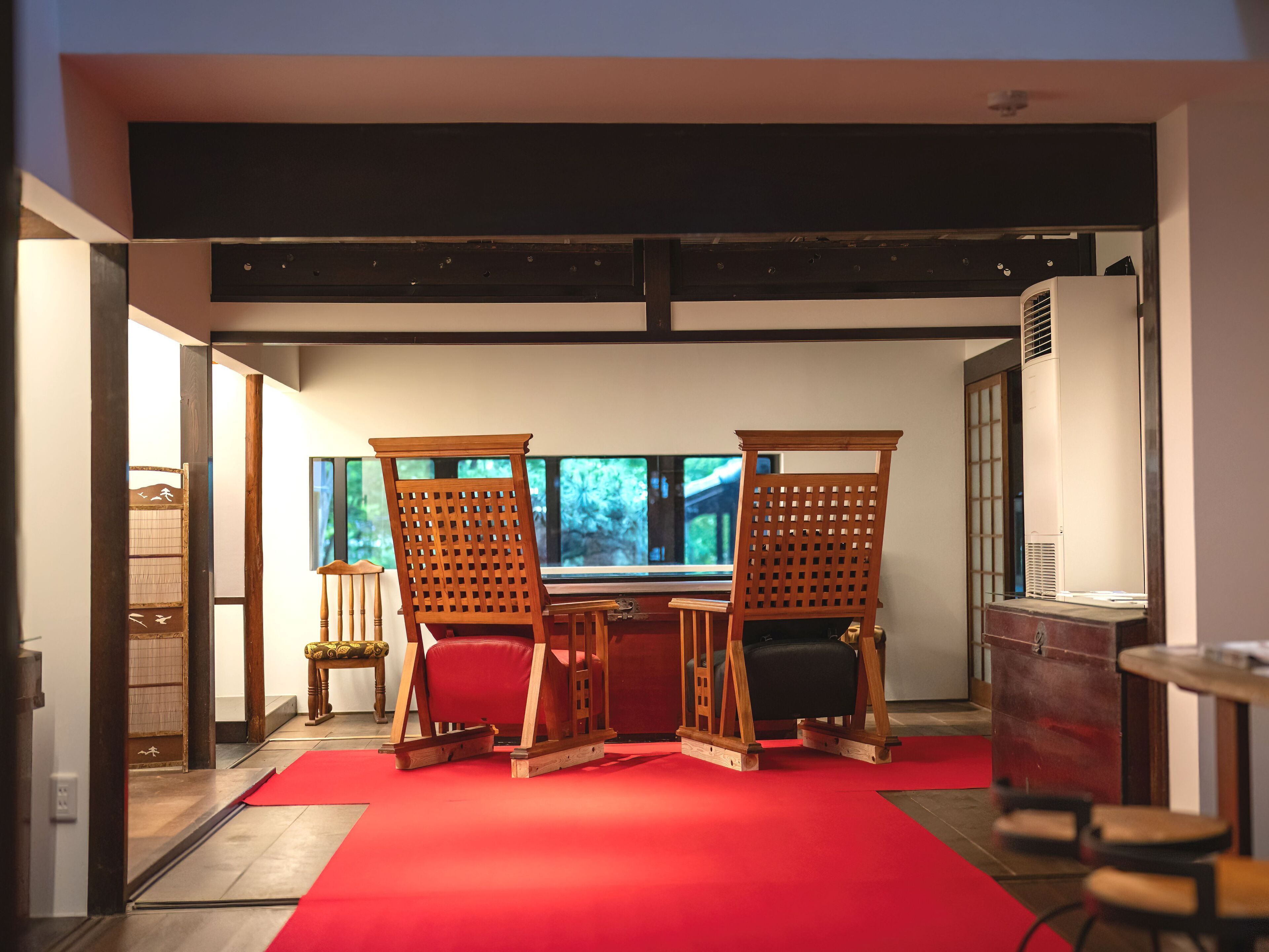 A room with a red carpet, wooden chairs, a table, and a large window with a view of greenery.