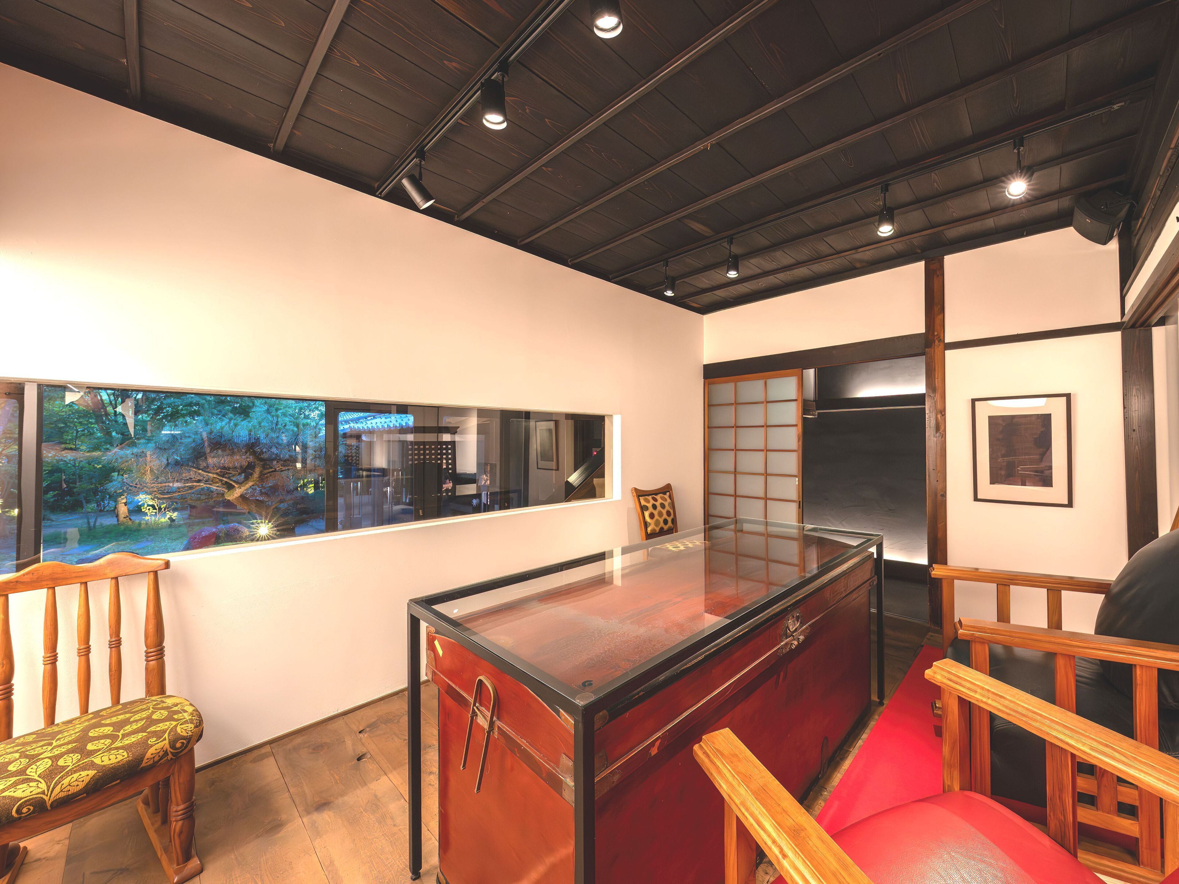 Una habitación con una mesa de vidrio, sillas de madera, un sofá rojo y un ventanal con vistas a un jardín.