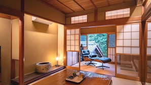 A traditional Japanese room with tatami flooring, a low wooden table with a tray, a cushioned chair, and sliding shoji doors leading to a view of a traditional building and trees. - Machiya Hotel Oyado Baison (Mino)