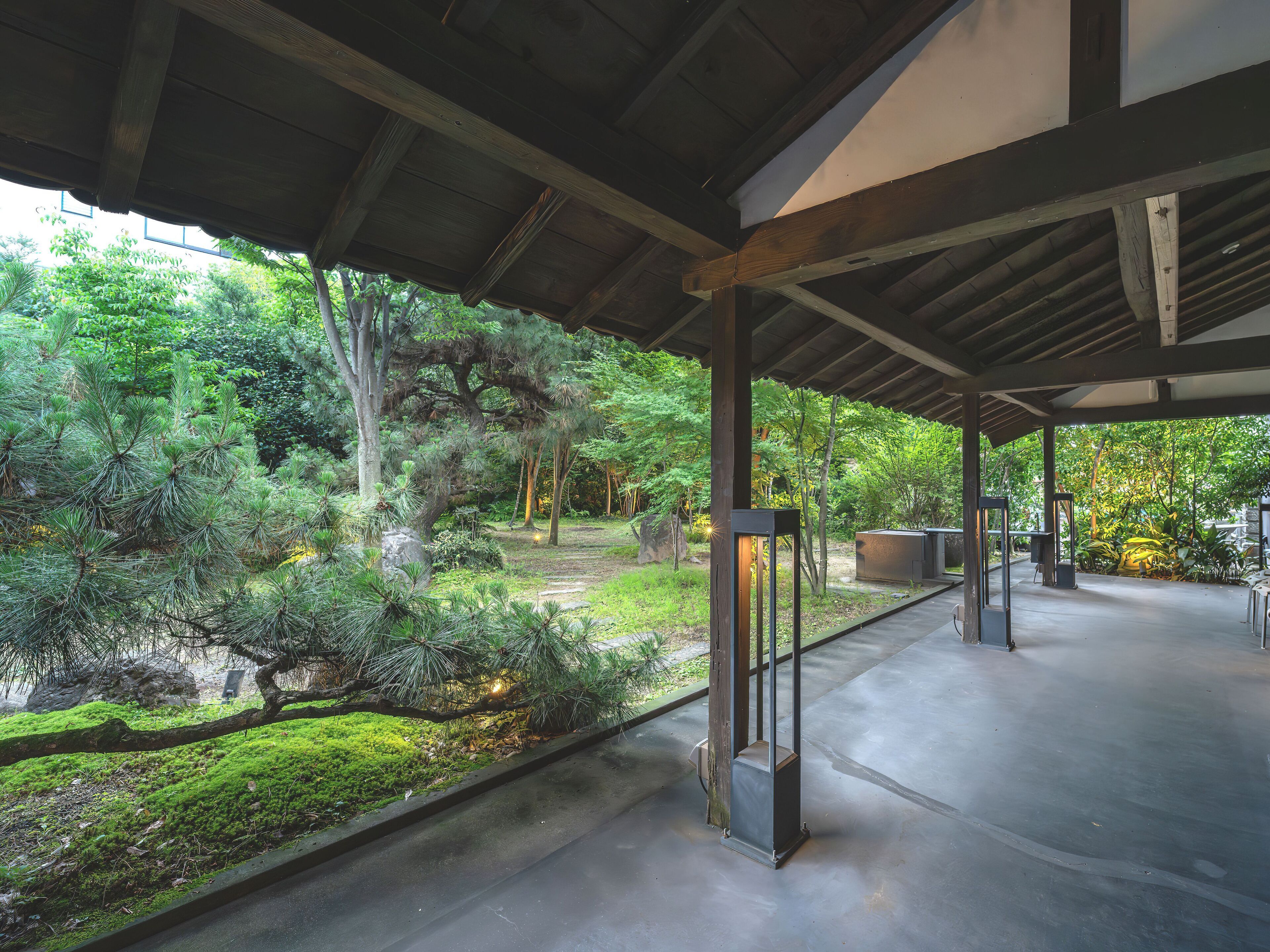 Um espaço coberto ao ar livre com piso de concreto, vigas de madeira e vista para um jardim com árvores e plantas.