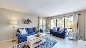 Ein Wohnzimmer mit zwei blauen Sofas, einem Glastisch und einem Teppich mit Muster.