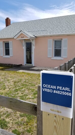 Une maison avec une plaque portant le numéro 205 au-dessus de la porte, une plaque bleue avec l’inscription « OCEAN PEARL VRBO #4825066 » devant, et une clôture en bois au premier plan.