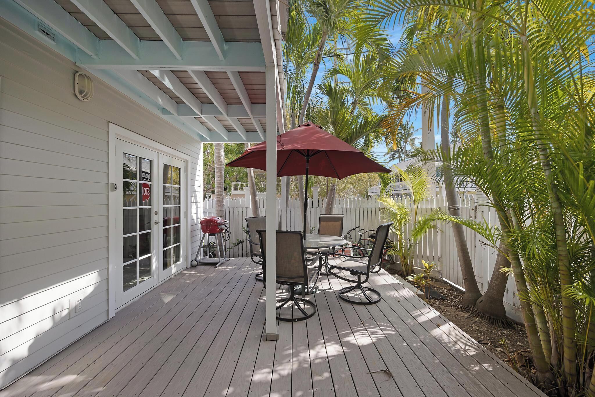 A covered patio with a table and chairs, a red umbrella, and a white fence with palm trees.