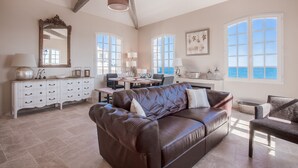 Ein großzügiger Wohnraum mit braunem Ledersofa, einem Essbereich mit Tisch und Stühlen, großen Fenstern mit Meerblick und einer weißen Kommode mit Schubladen.