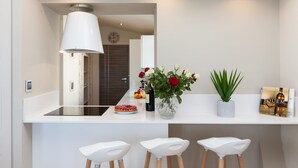 Eine moderne Küche mit weißer Arbeitsplatte, drei weißen Barhocker, einer großen weißen Pendelleuchte, einer Vase mit roten Rosen, einem Blumentopf und einer Zeitschrift auf der Arbeitsfläche.