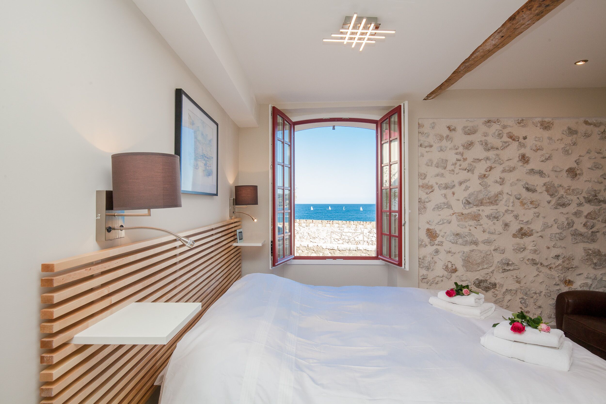 Una camera da letto con testiera in legno a listoni, un comodino, una lampada, un quadro incorniciato, vista sul mare da una finestra aperta e una parete in pietra.