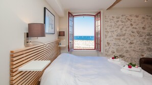 Ein Schlafzimmer mit einem Holz-Schranken-Kopfstück, einem Nachttisch, einer Lampe, einem gerahmten Bild, Blick aufs Meer durch ein offenes Fenster und einer Steinwand.