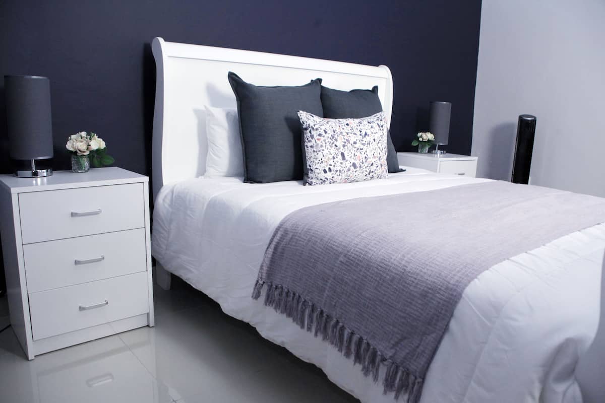 Ein modernes Schlafzimmer mit einem weißen Bett, dunklen Kissen, einer grauen Tagesdecke und einem Nachttisch mit Lampe und Blumenvase.