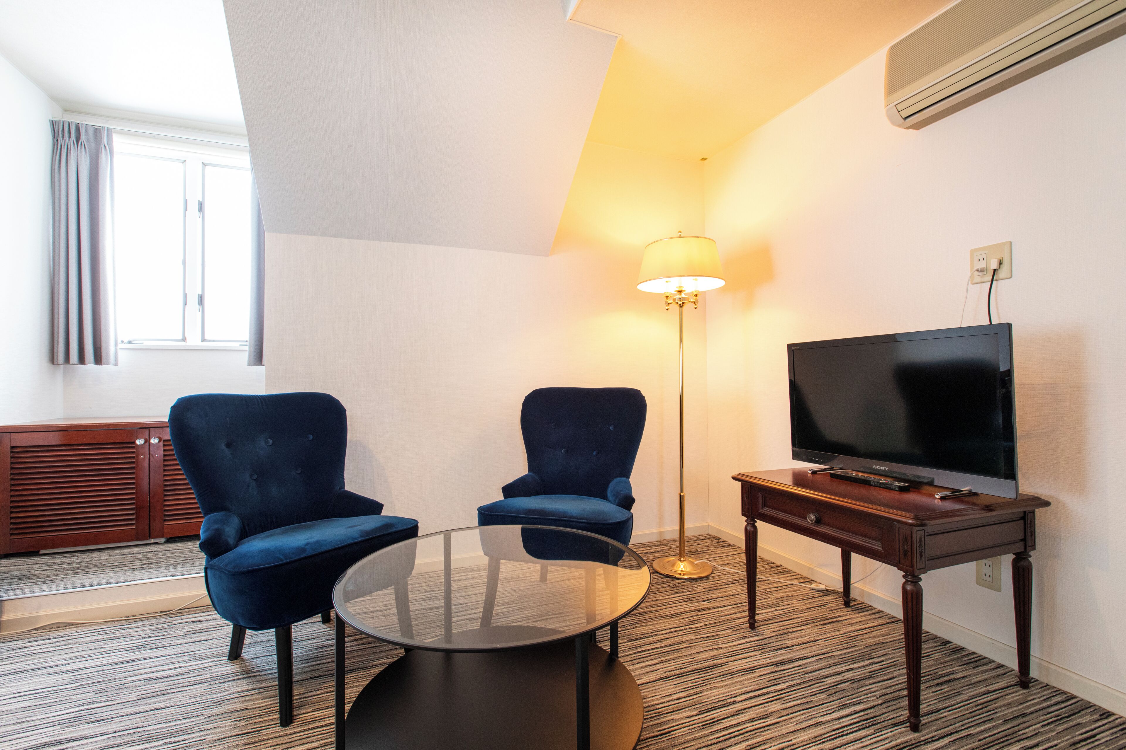 A hotel room with a TV, two blue armchairs, a glass coffee table, a floor lamp, and a wooden cabinet.