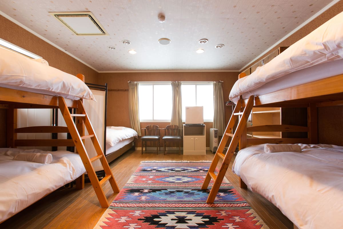 A dormitory room with bunk beds, a desk, chairs, and a patterned rug.