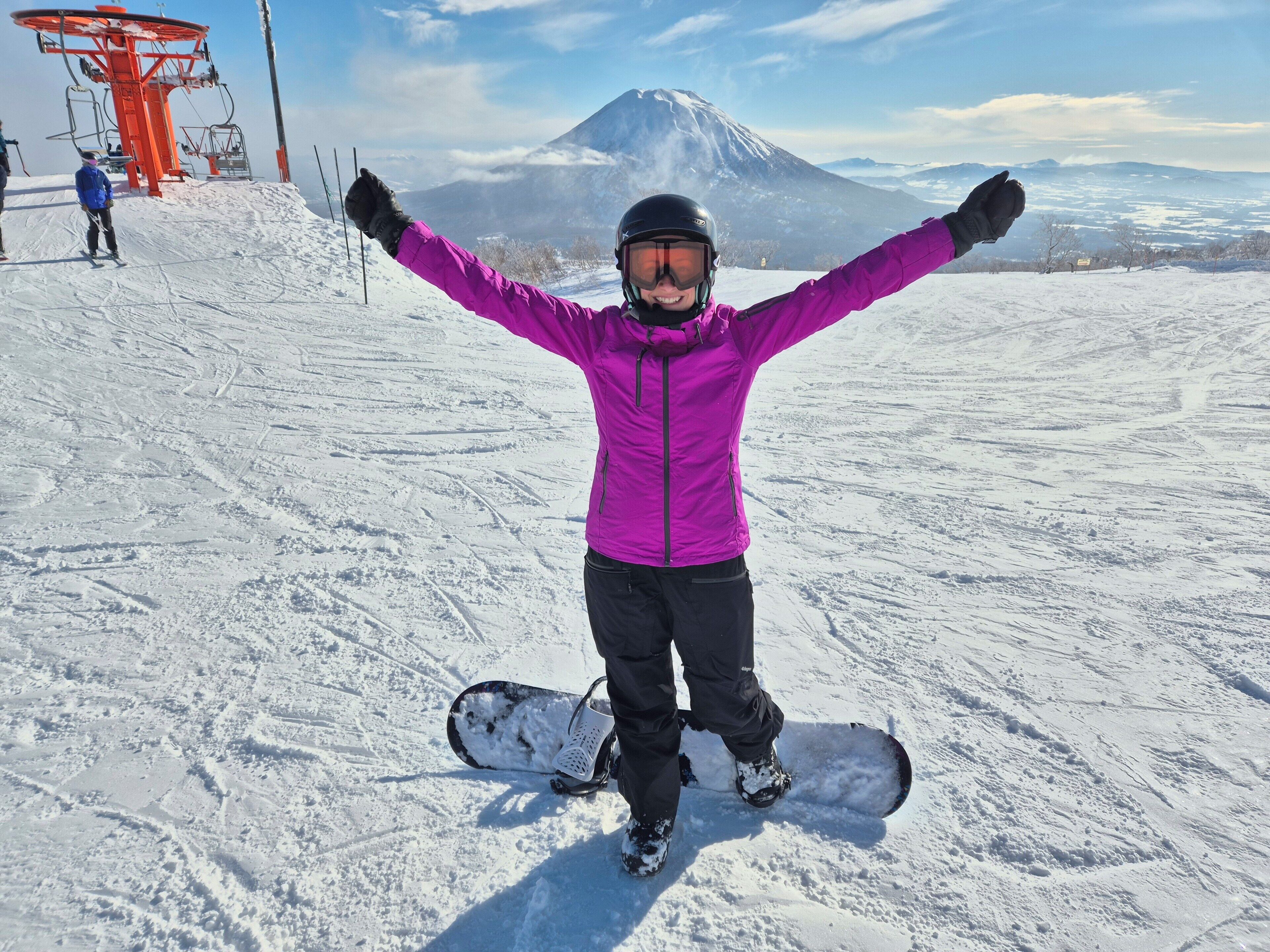 A person with arms raised, standing on a snowy slope with a snowboard, a ski lift in the background, and a mountain in the distance.