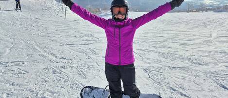 A person with arms raised, standing on a snowy slope with a snowboard, a ski lift in the background, and a mountain in the distance.