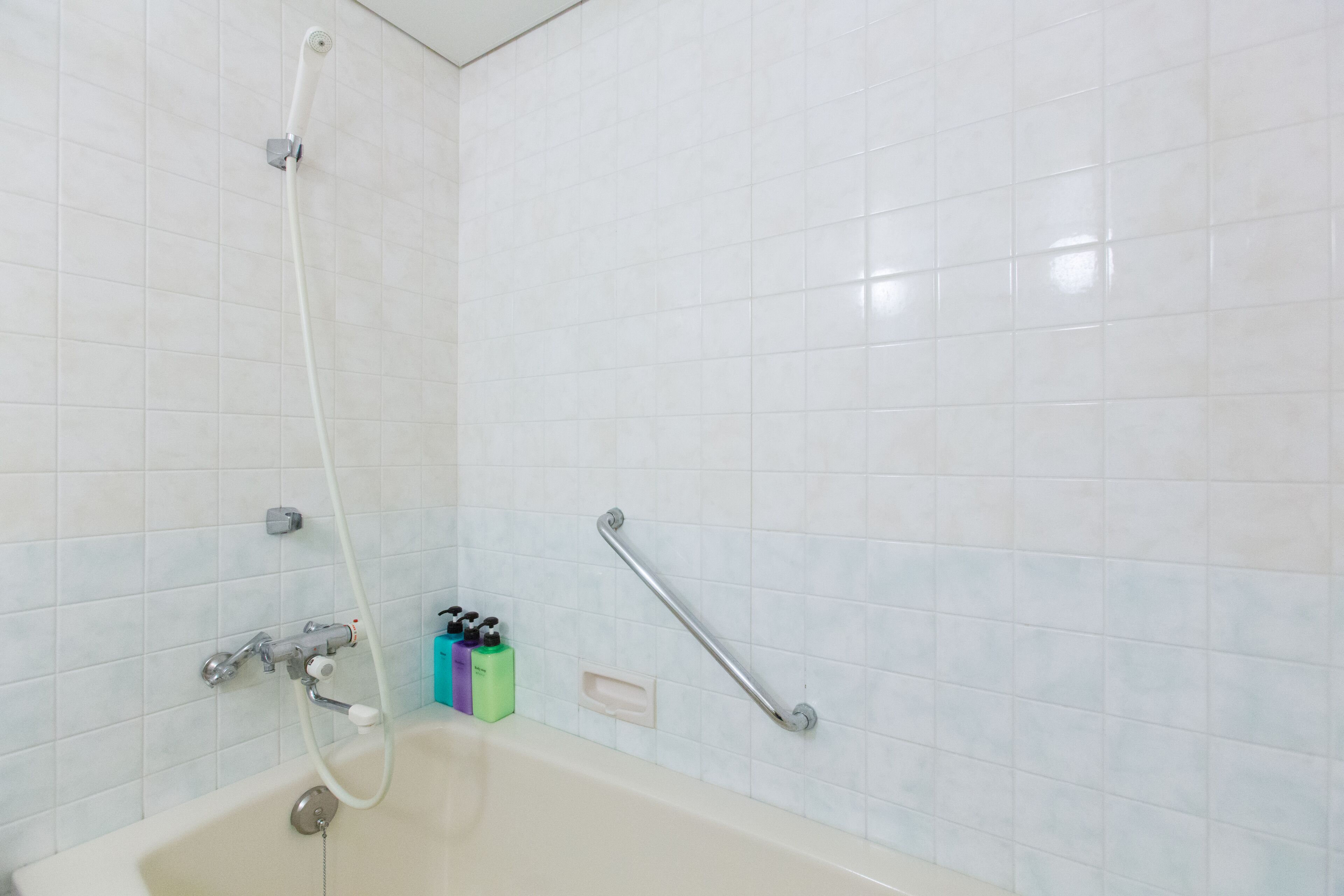 Un baño con paredes de azulejos blancos, una bañera, un cabezal de ducha manual y una barra de apoyo.