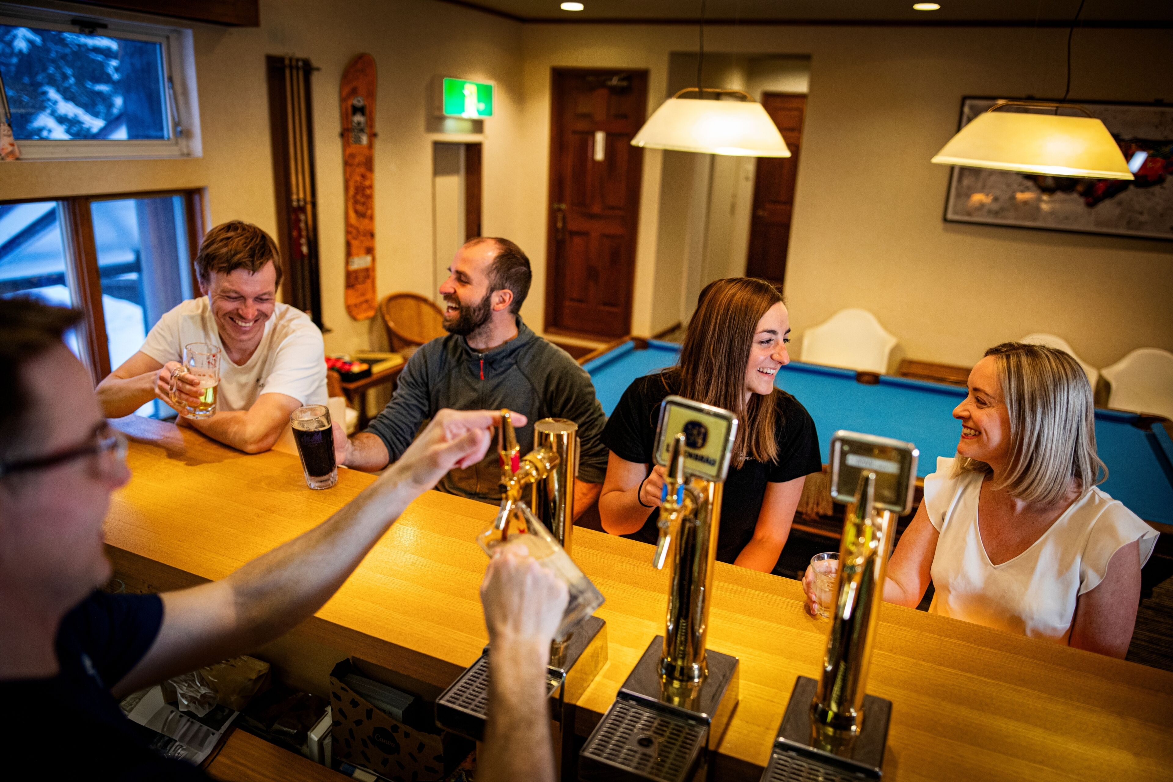 A group of people at a bar with multiple taps, a pool table, and a wooden table.