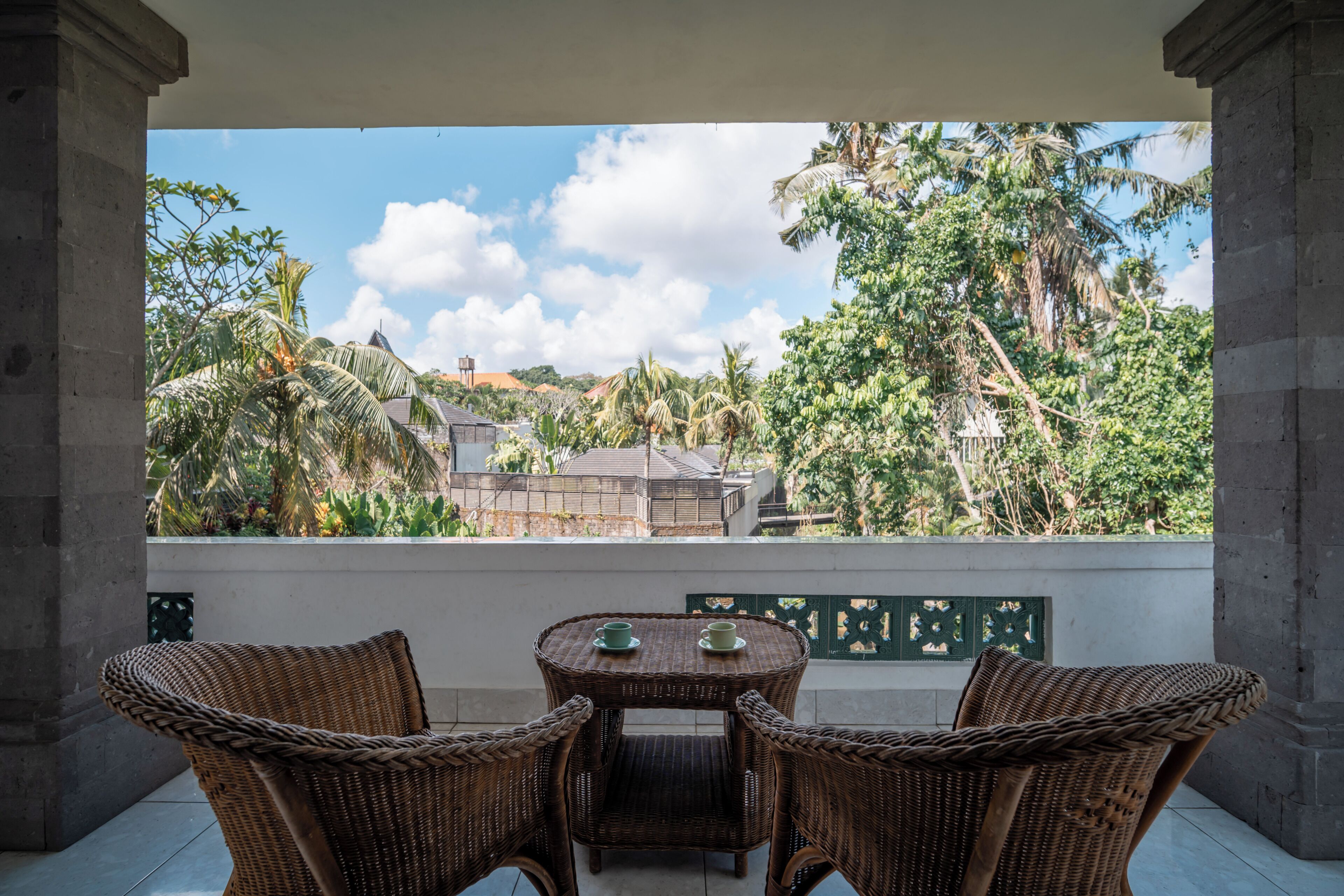 A balcony with wicker furniture, a table, and a view of tropical trees and buildings.