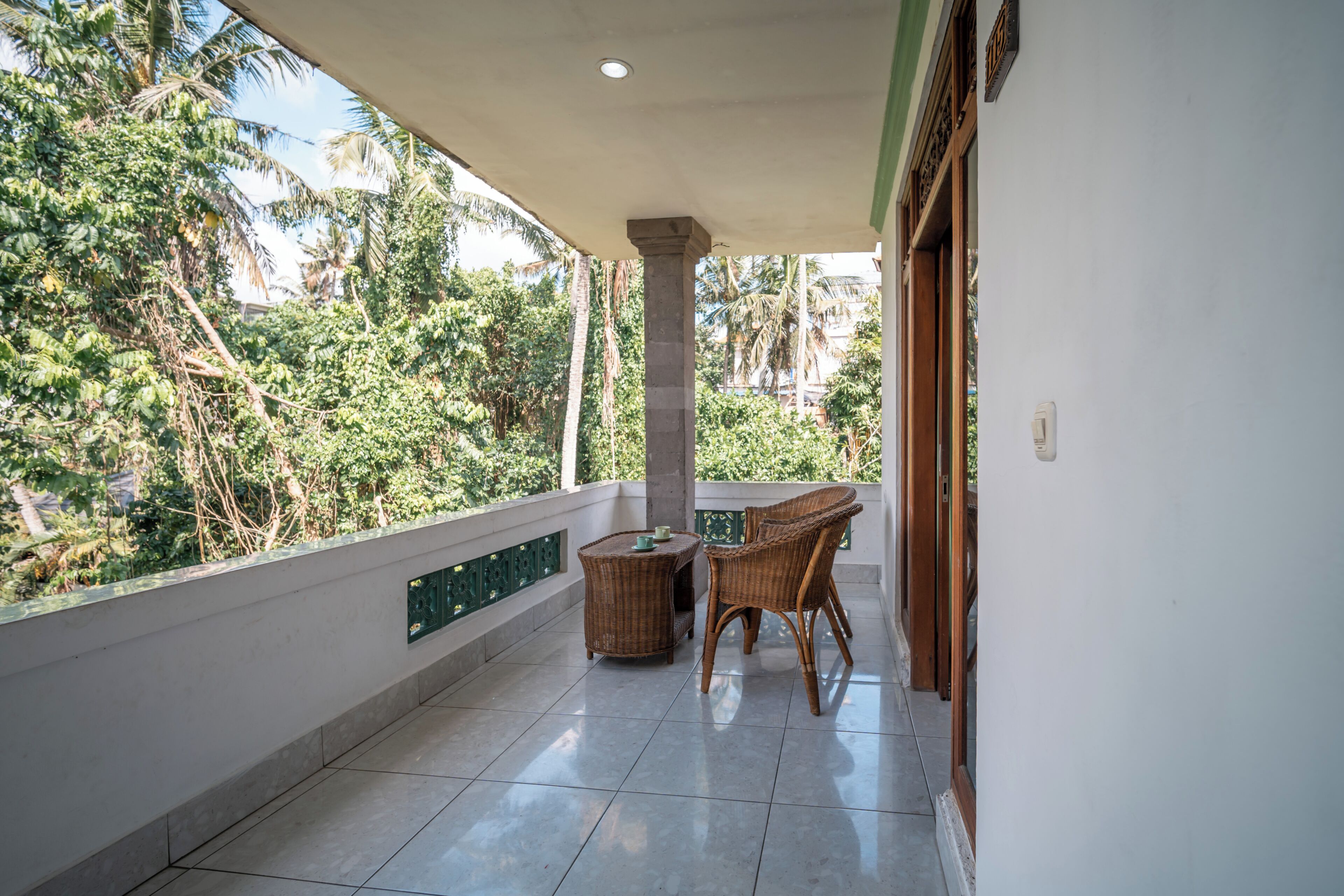 A balcony with a tiled floor, a wicker table, two wicker chairs, and a view of greenery.