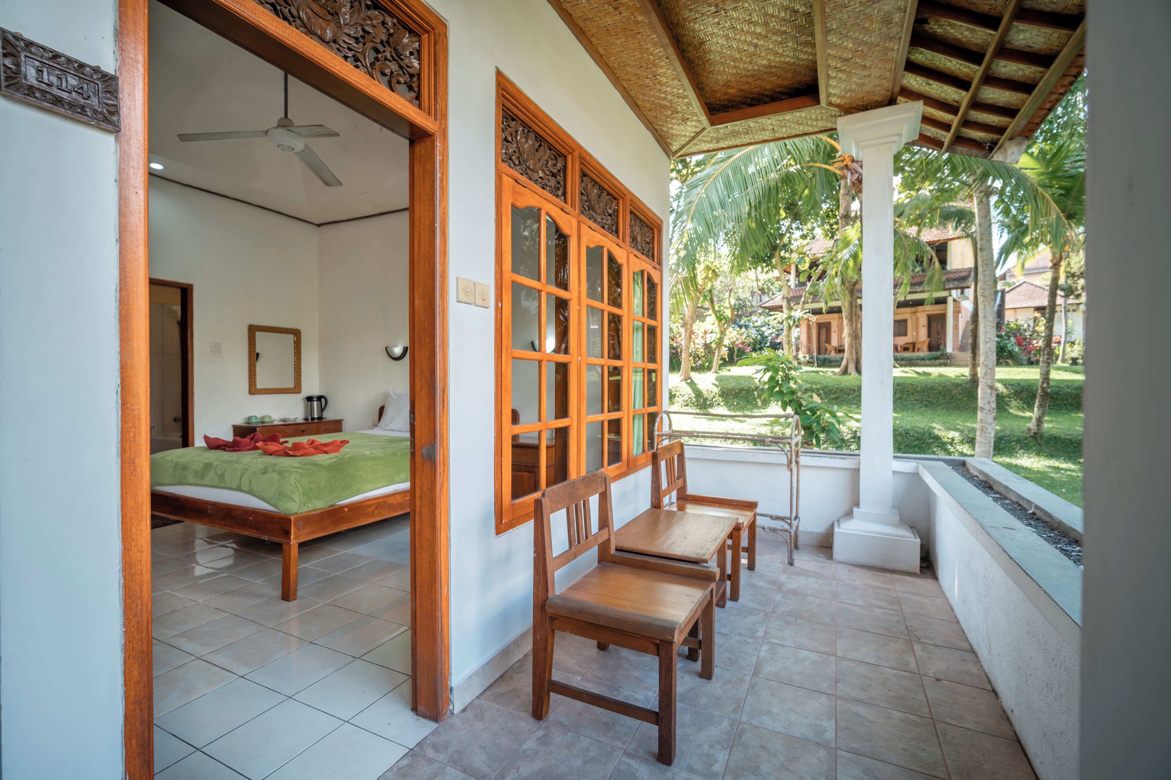 Quarto com cama, escrivaninha e cadeira, janela com vista para um jardim e ventilador de teto.