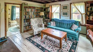 A cozy living room with a wooden coffee table, a blue sofa, a floral rug, and a bookshelf filled with books. - Chester Farmhouse on 100 Acres, 15 Min to Okemo! (Chester)