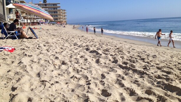 A beach with people walking and sitting under colorful umbrellas. There is a building with multiple balconies and a palm tree in the background. - Perfect Laguna Beach Vacation-ocean views/beach across street/town 1 block (Laguna Beach)