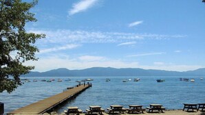 Ein See mit Sandstrand, Picknicktischen und Liegestühlen, einer Holzstege und Booten auf dem Wasser.