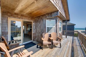 Eine Holzterrasse mit zwei Adirondack-Stühlen, einem Glastür-Eingang und Blick auf den Ozean.