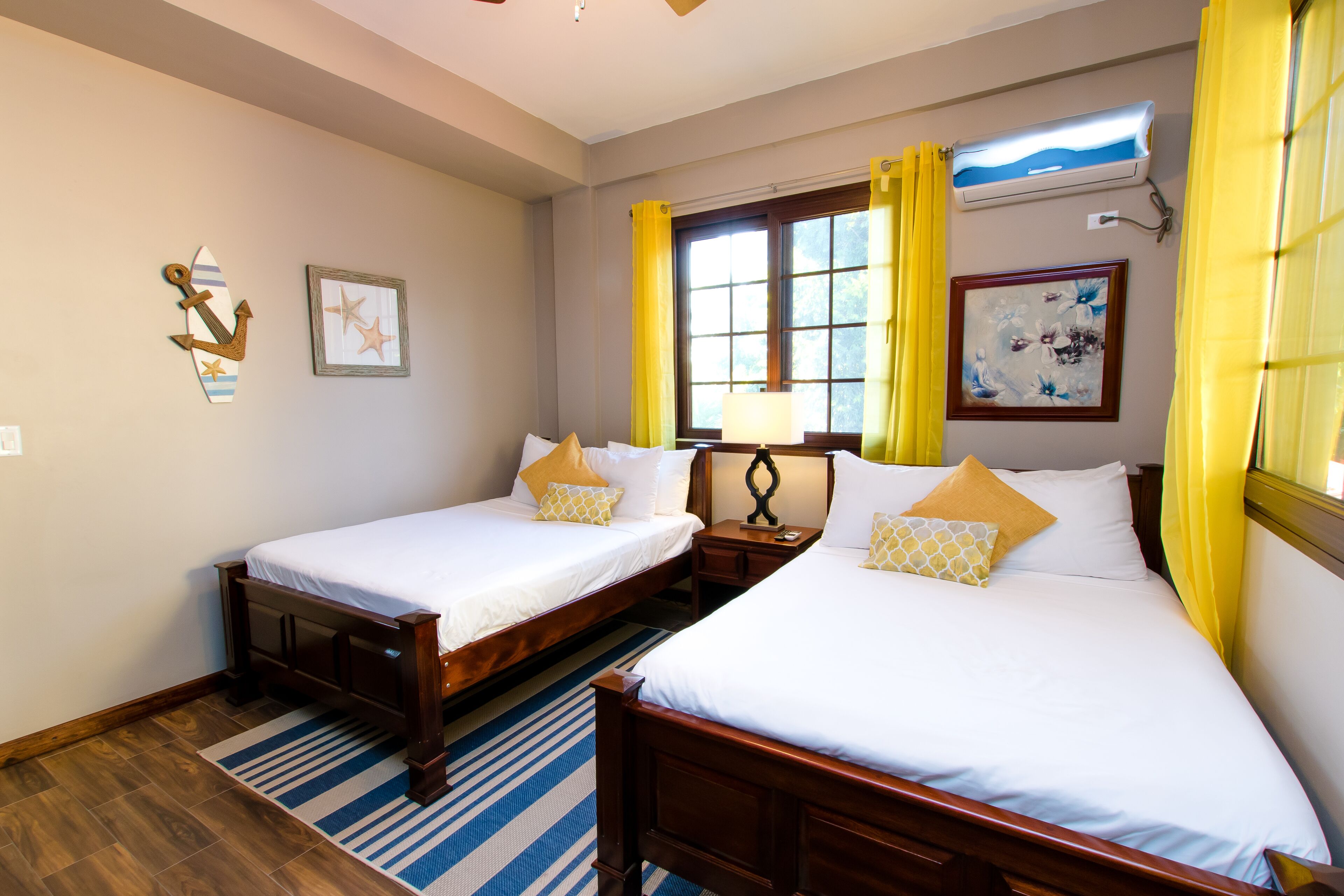 Habitación de hotel con dos camas, cabeceros de madera, una ventana con cortinas amarillas, un cuadro enmarcado y un aire acondicionado montado en la pared.