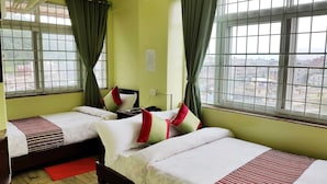 A hotel room with two single beds, a bedside table, a fan, and a window with a view of buildings. - Hotel Arhant Inn (Pokhara)