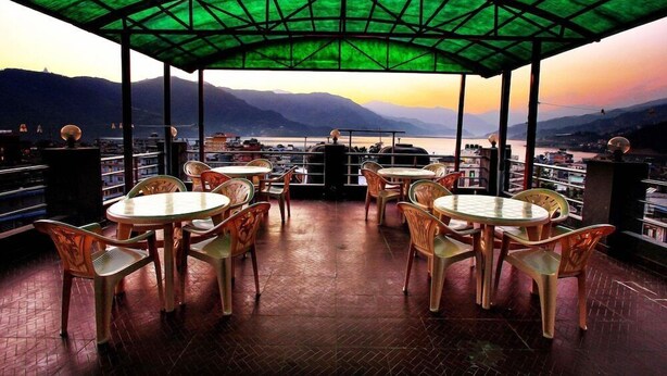 An outdoor terrace with round tables and chairs, a green roof, and a view of mountains and a lake. - Hotel Arhant Inn (Pokhara)