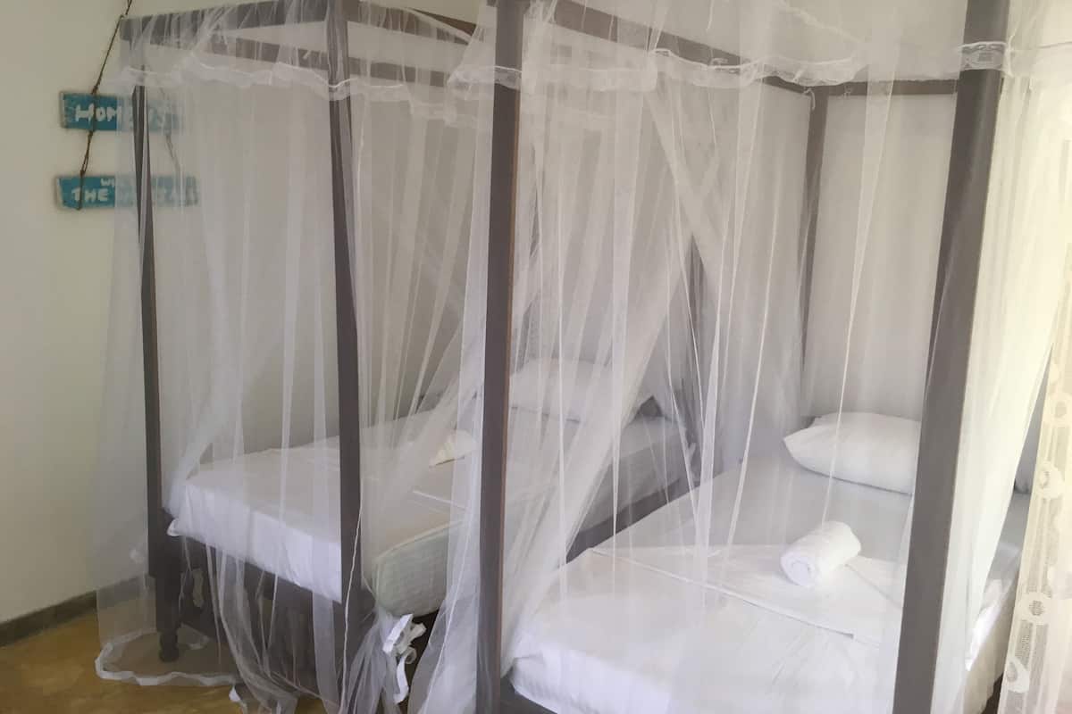 A room with two single beds covered with white sheets and a mosquito net. There is a sign on the wall that reads "Home."