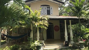 A two-story building with a beige facade, a tiled roof, and a covered porch with columns. There is a hammock and a potted plant in the foreground, and a sign reading "Villa Coconut" on the building. - Villa Coconut - Hostel (Weligama)