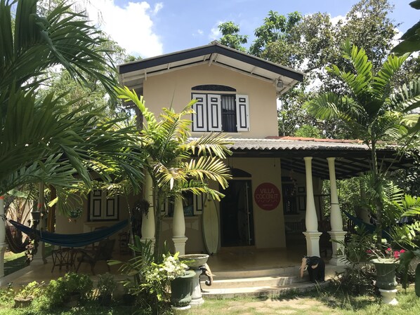 A two-story building with a beige facade, a tiled roof, and a covered porch with columns. There is a hammock and a potted plant in the foreground, and a sign reading "Villa Coconut" on the building. - Villa Coconut - Hostel (Weligama)