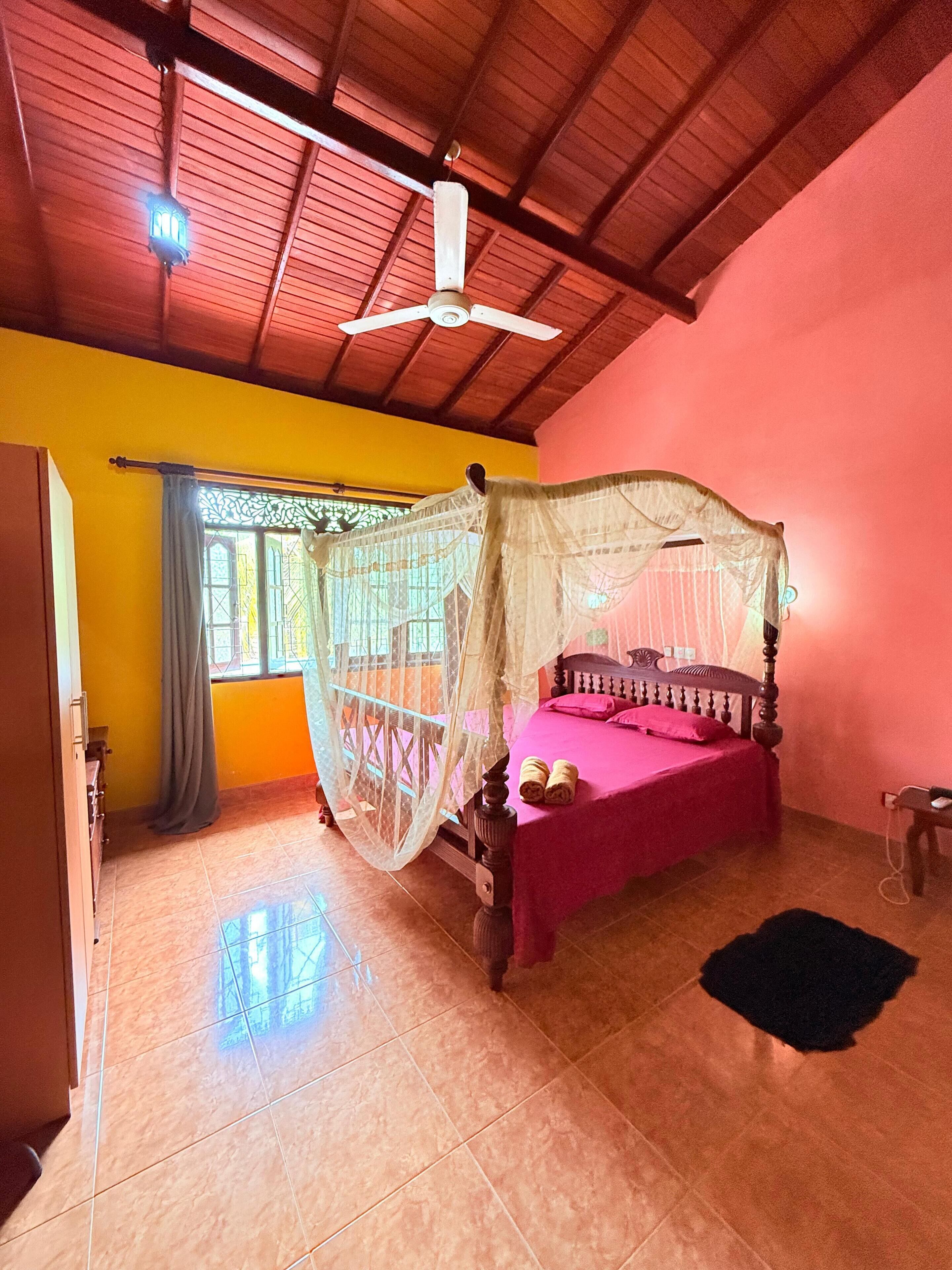 Een kamer met een bed, een plafondventilator, een raam met gordijnen en een betegelde vloer.