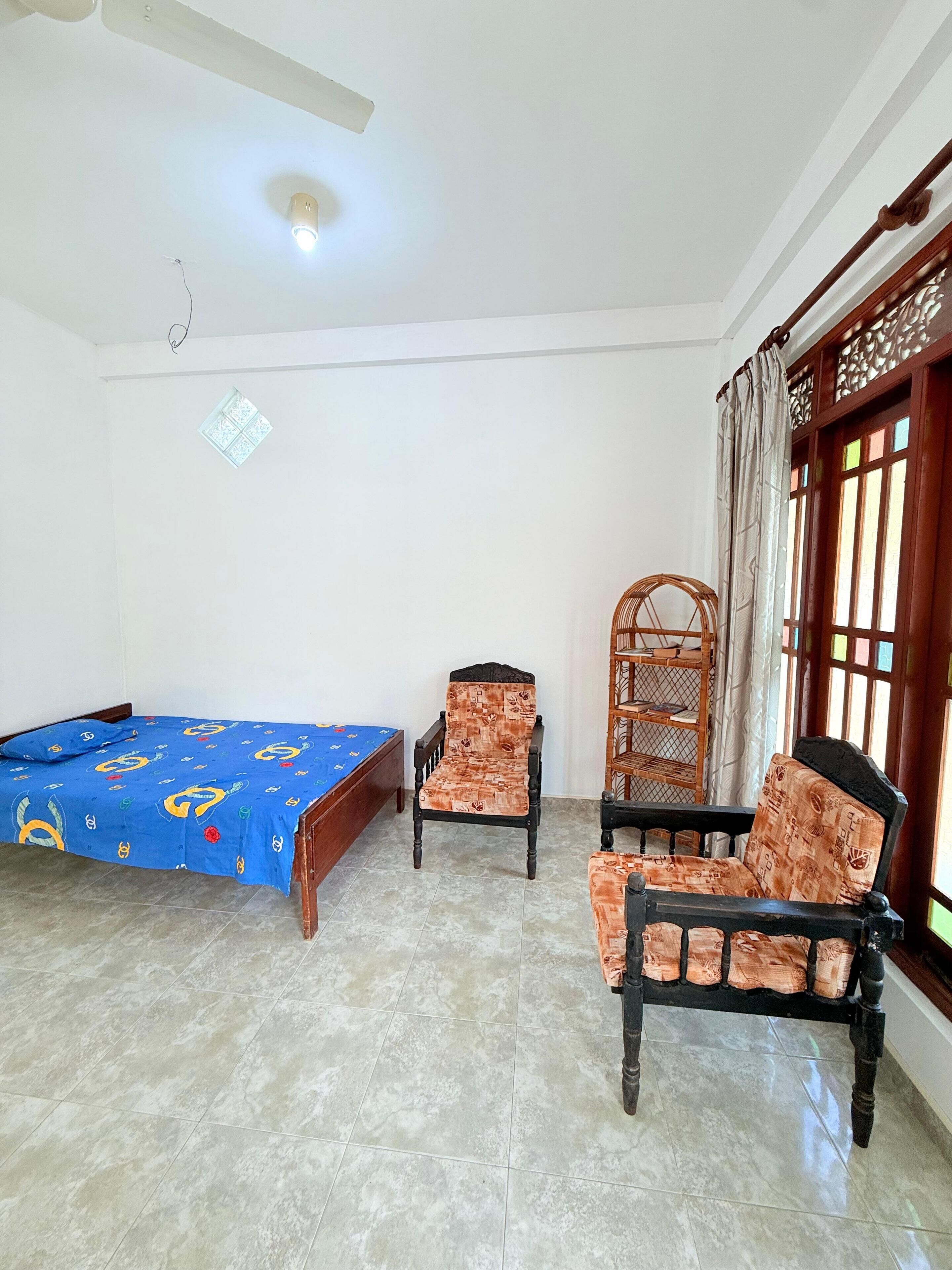 A room with a bed, two chairs, a window with curtains, and a ceiling fan.