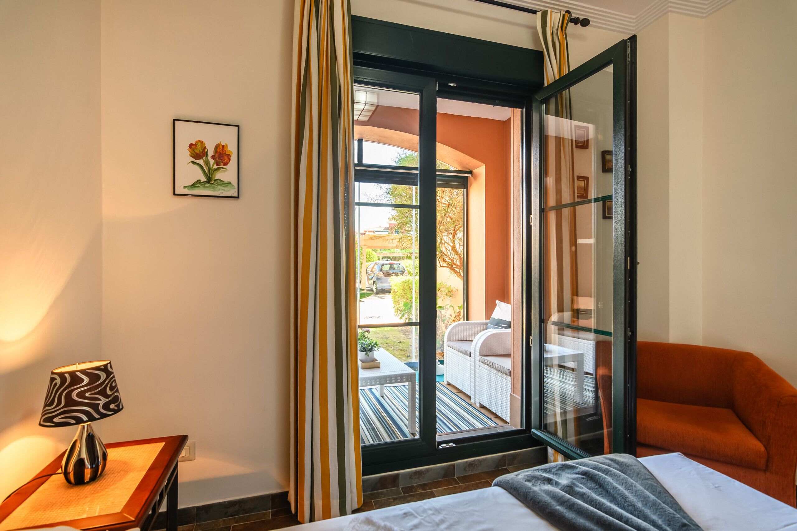 A hotel room with a bed, a chair, a table with a lamp, a sliding glass door to an outdoor area with a table and chairs, and a framed picture on the wall.