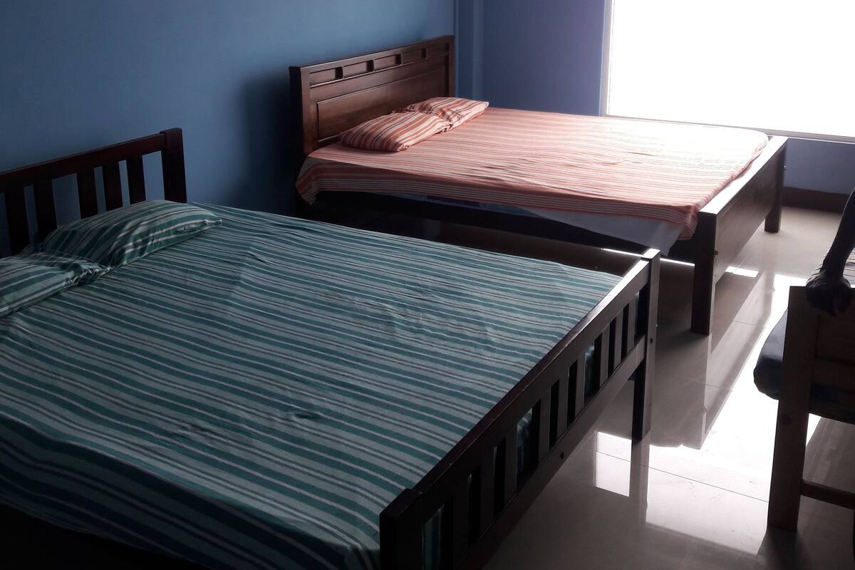 A room with two single beds, a wooden headboard, and a window with blinds.