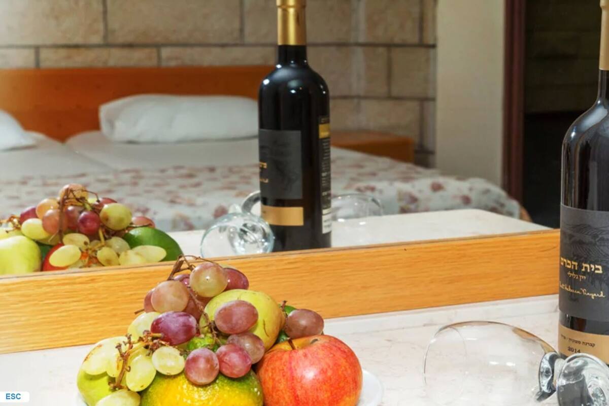 Quarto de hotel com uma cama, uma mesinha de cabeceira com uma garrafa de vinho, uma cesta de frutas com uvas e uma maçã, e um espelho refletindo o quarto.