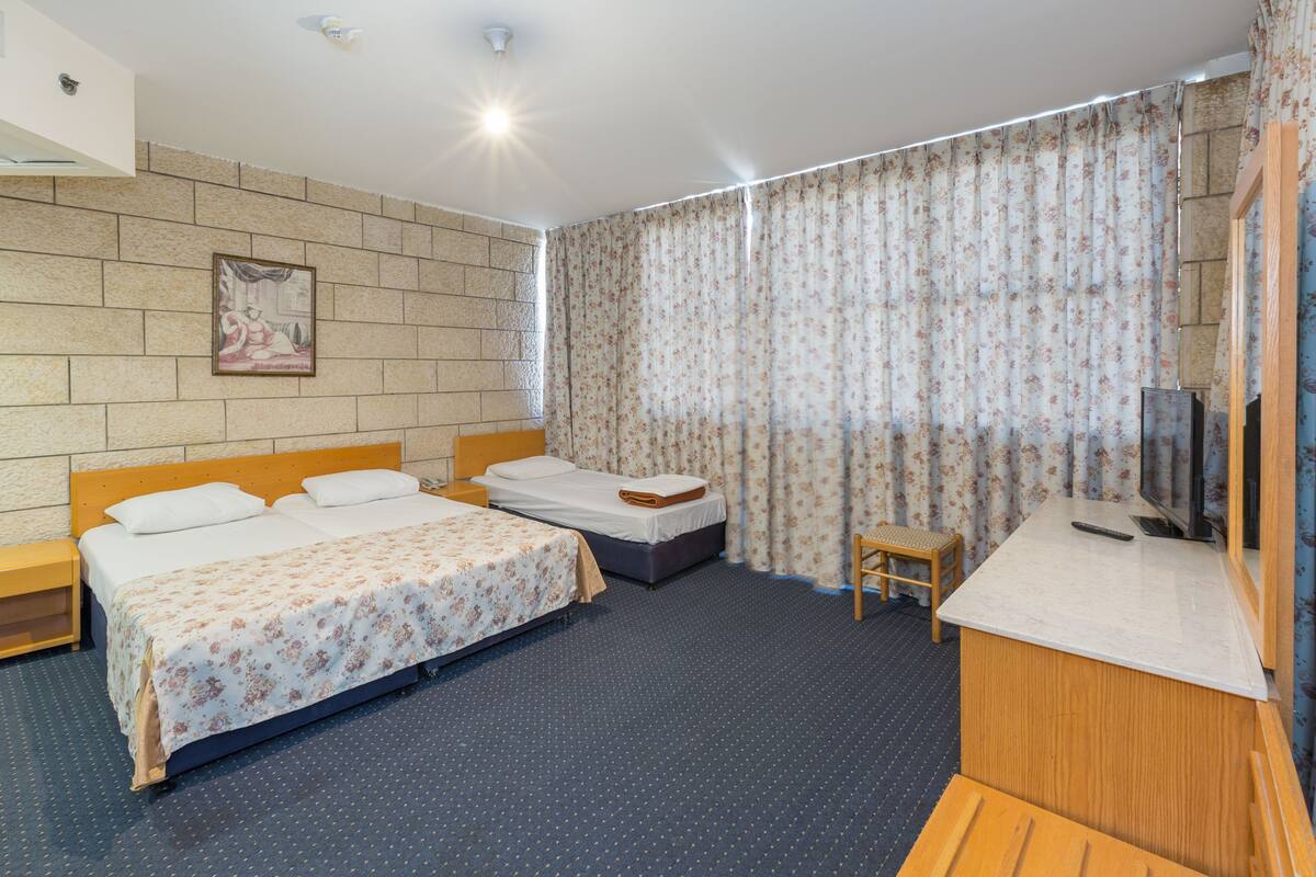 Quarto de hotel com duas camas, uma pequena escrivaninha, televisão e janela com cortinas.
