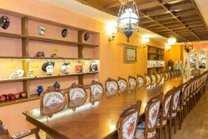 Uma longa mesa de madeira com cadeiras decoradas, rodeada por estantes com máscaras e outros objetos decorativos.