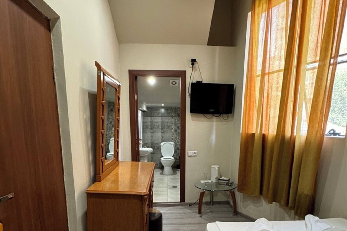 Ein Hotelzimmer mit einem Holztisch, einem Bett mit ordentlich gefalteten Handtüchern, einem an der Wand montierten Fernseher und einem durch eine offene Tür einsehbaren Badezimmer.