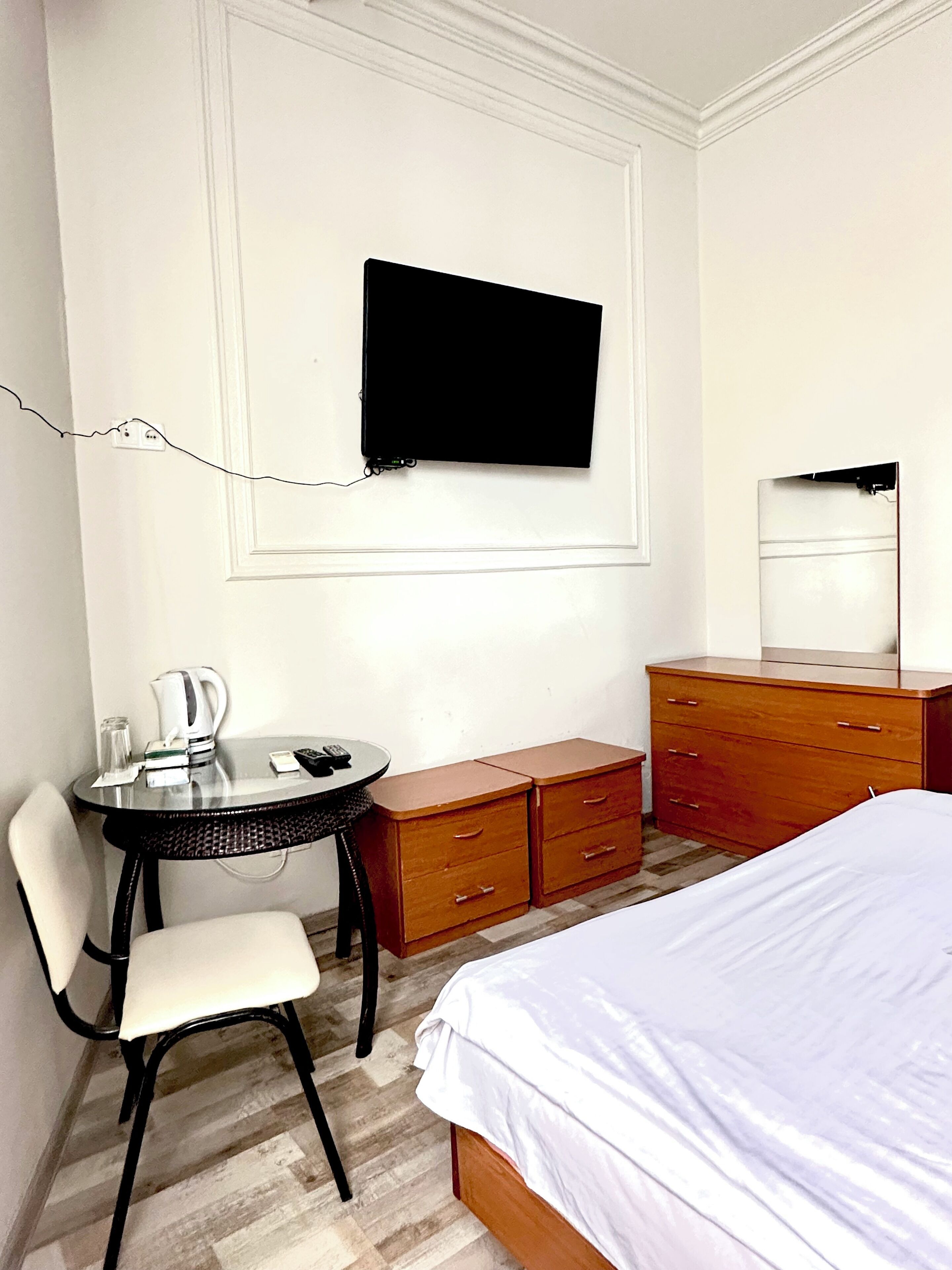 A hotel room with a bed, a desk with a chair, a dresser, a television mounted on the wall, and a small table with a kettle and remote controls.