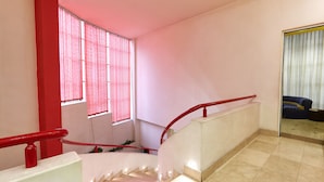A staircase with red railings and a handrail, leading to a landing with a view of a room with a blue sofa and a window with vertical blinds. - Bella Hotel (Yerevan)