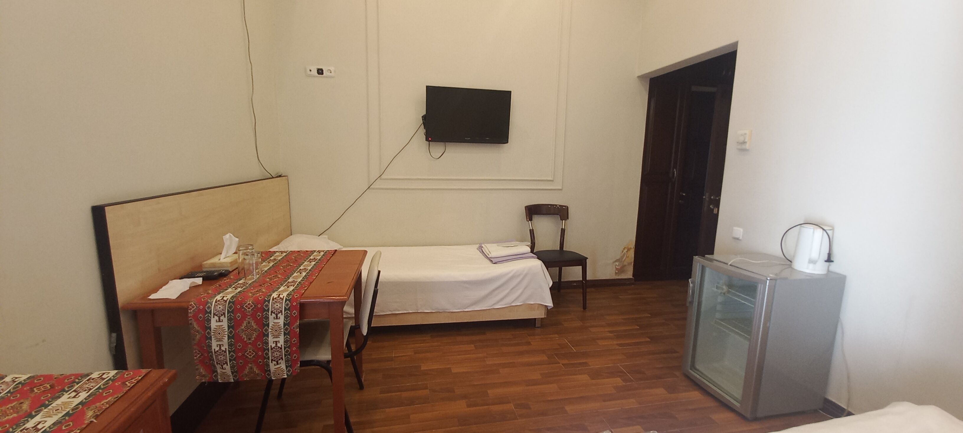 A small, well-lit room with a wooden floor, a bed with a white bedspread, a desk with a patterned tablecloth, a chair, a television mounted on the wall, and a small refrigerator.