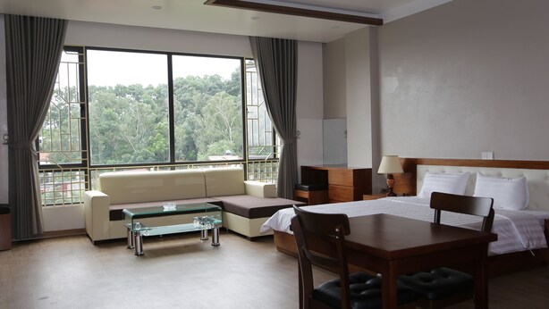 A hotel room with a bed, a desk, a sofa, and a large window offering a view of trees.