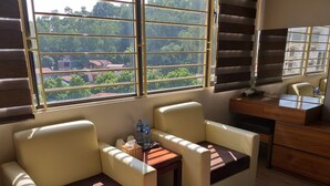 A room with two cushioned chairs, a small table with water bottles, and a desk with a mirror. Large windows provide a view of buildings and trees outside.